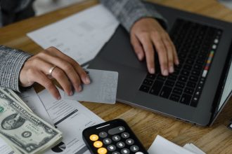 Pessoa usa cartão de crédito ao lado de dinheiro, calculadora e laptop em uma mesa.
