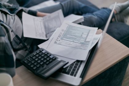 Pessoa usa um notebook e uma calculadora para organizar documentos financeiros em casa.